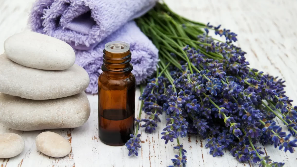 An image of a rustic white table with massage stones, massage oil, lavender flowers, and purple towels rolled up next to it.